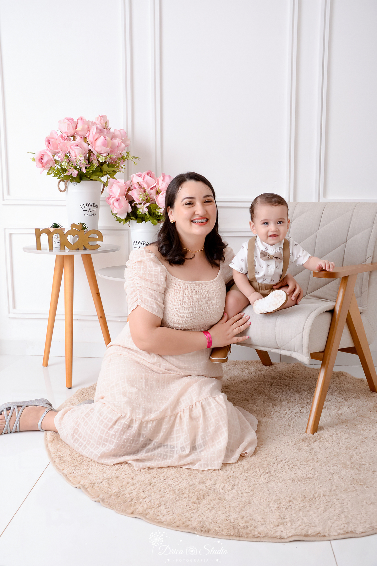 drica studio, ensaio fotografico, xinguara, dia das mães, mãe sentado no chão segurando bebê sentdo em poltrona, vestindo bege, arranjo de flores, fundo branco, short com suspensório e camisa branca.