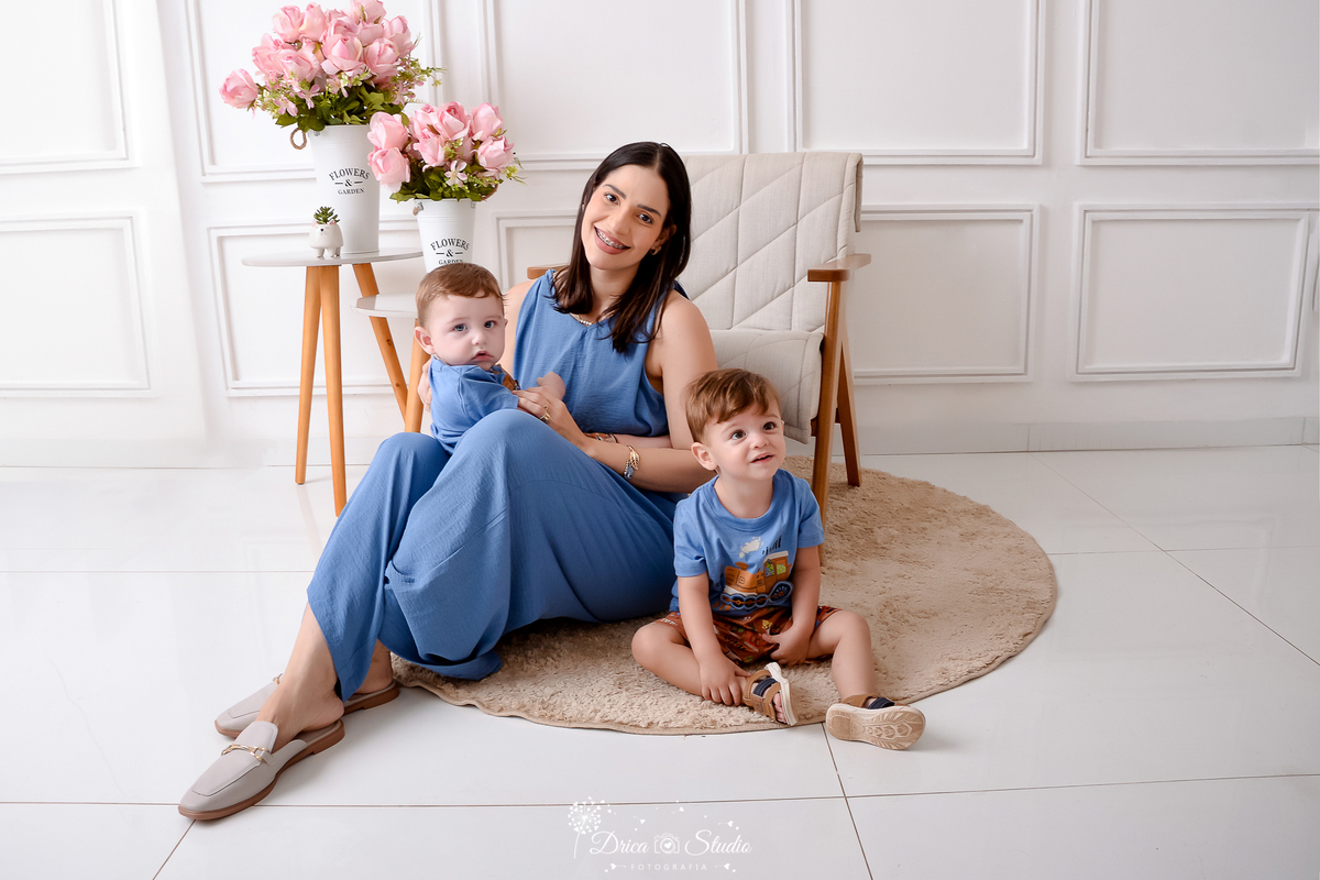 drica studio, ensaio fotografico, xinguara, dia das mães, mãe sentanda no chão com os dois filhos, maquinho longo azul, camisetas azuis, fundo branco com boiserie, flores rosa, carpete redondo bege.