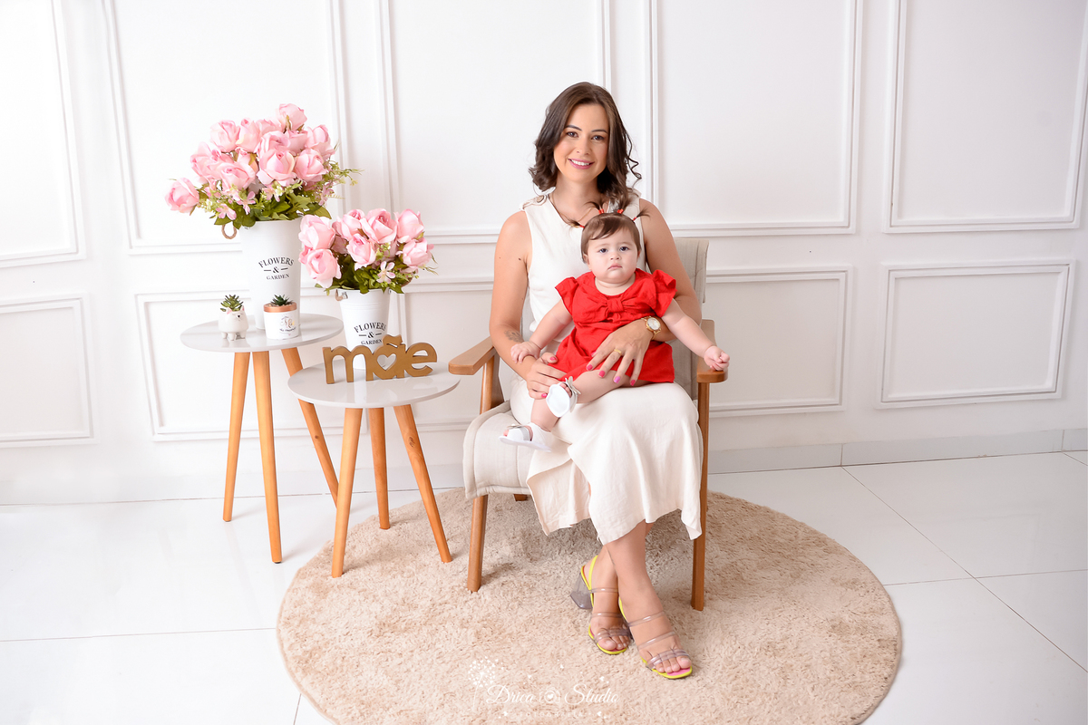 drica studio, ensaio fotografico, xinguara, dia das mães, mãe sentada com bebê no colo, vestido vermelho e vestido branco, flores rosa, vaso branco, funco branco com boiserie, mesinha de canto, carpete redondo bege