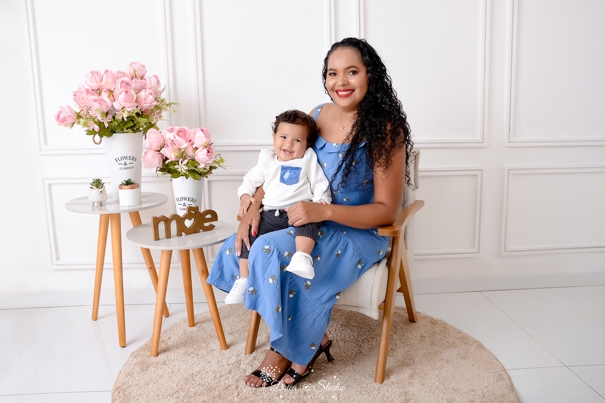 drica studio, ensaio fotografico, xinguara, dia das mães, mãe sentada com filho pequeno no colo, camiseta branca, vestido azul, flores rosa e vaso branco, mesinha de canto, poltrona de madeira e estofado bege claro, funco branco com boiserie