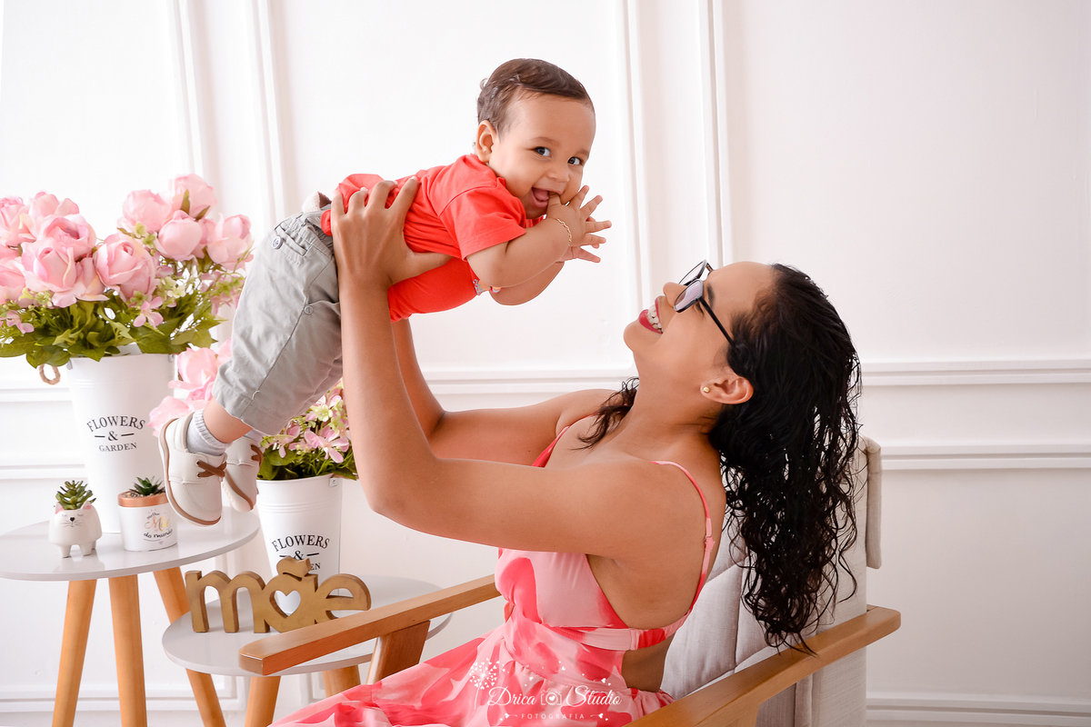 drica studio, ensaio fotografico, xinguara, dia das mães, mãe segurando bebê no alto, bebê vestindo short cor areia e camiseta vermelha, vestindo floral, fundo branco com boiserie, flores rosas e vaso branco,