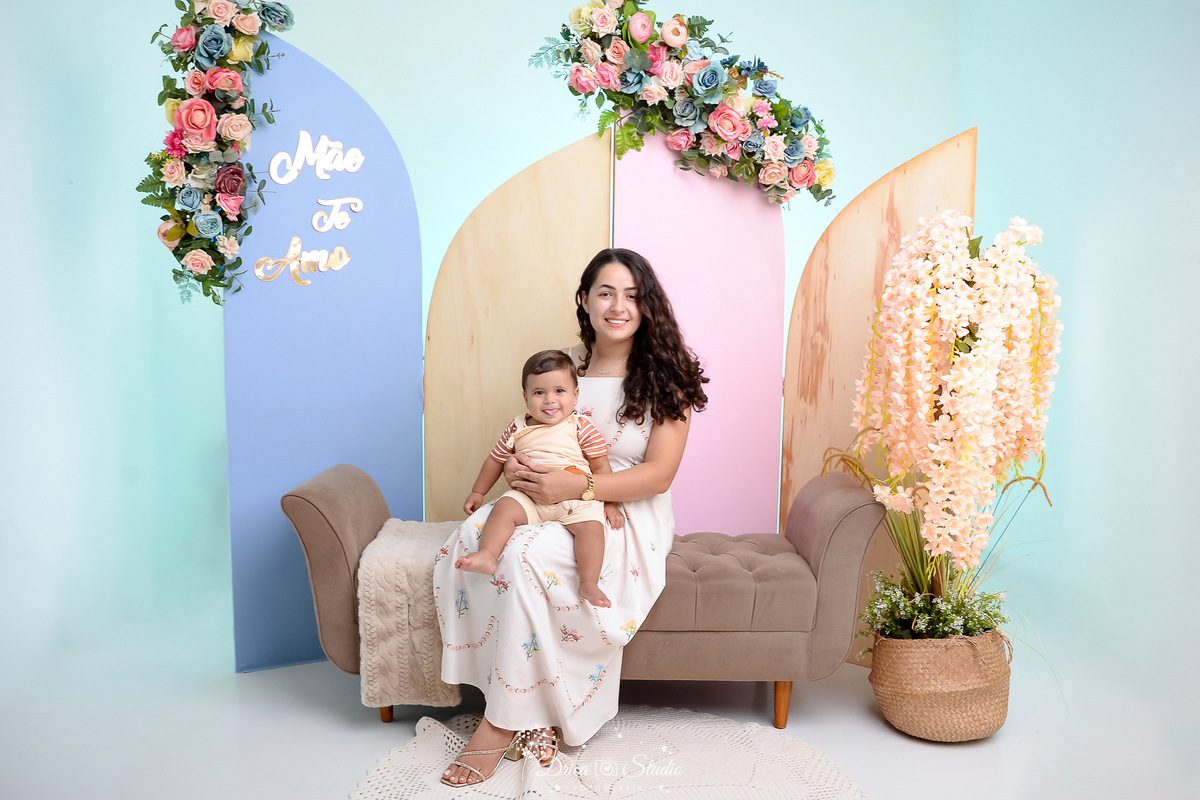 drica studio, ensaio fotografico, xinguara, dia das mães, mãe sentada em divam marrom com filho no colo, fundo azul, painel azul e rosa, arranjo floral, vestido branco e maquinho bege desenho de um leãozinho