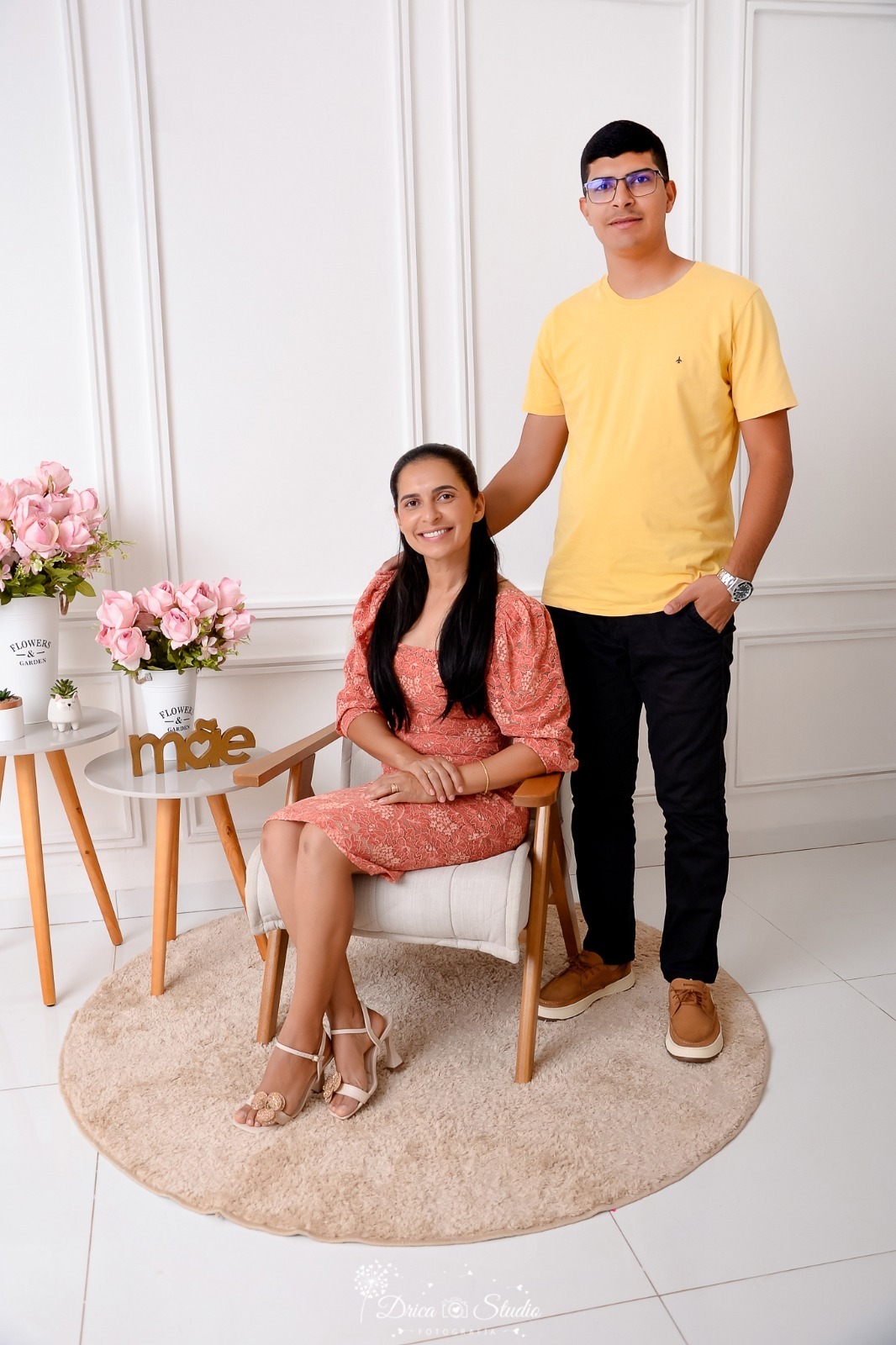 drica studio, ensaio fotografico, xinguara, dia das mães, vestindo longo cor damasco, calça preta e camisa amarela, mãe sentada em poltrona e filho em pé ao lado, mesinha de canto, fundo branco com boiserie, vaso branco com flores rosa