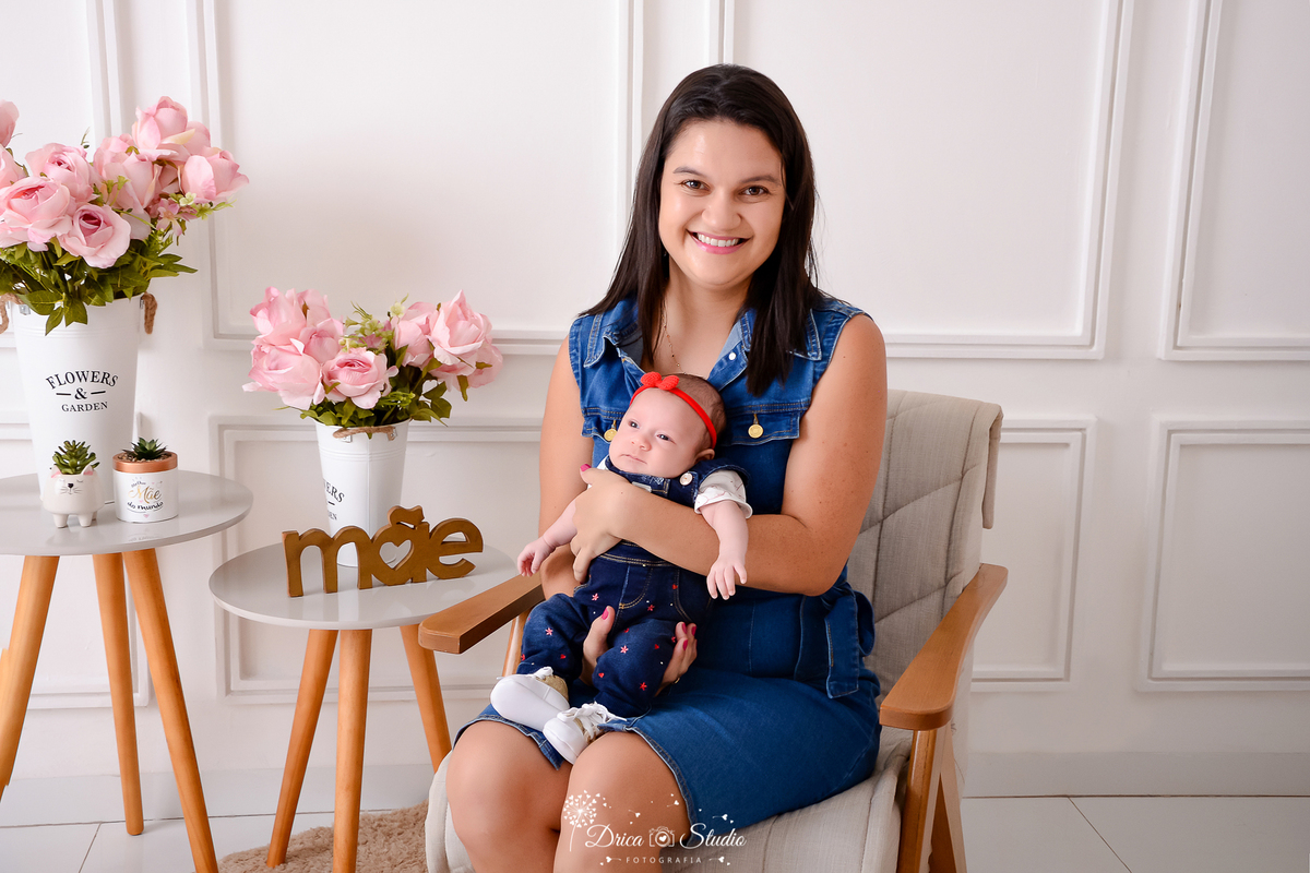 drica studio, ensaio fotografico, xinguara, dia das mães, mãe segurando bebê de 1 mês, vestido jeans, maquinho jeans e blusa branca, fundo branco com boiserie, flores rosa, vaso branco, mesinha de canto, estofado cinza claro