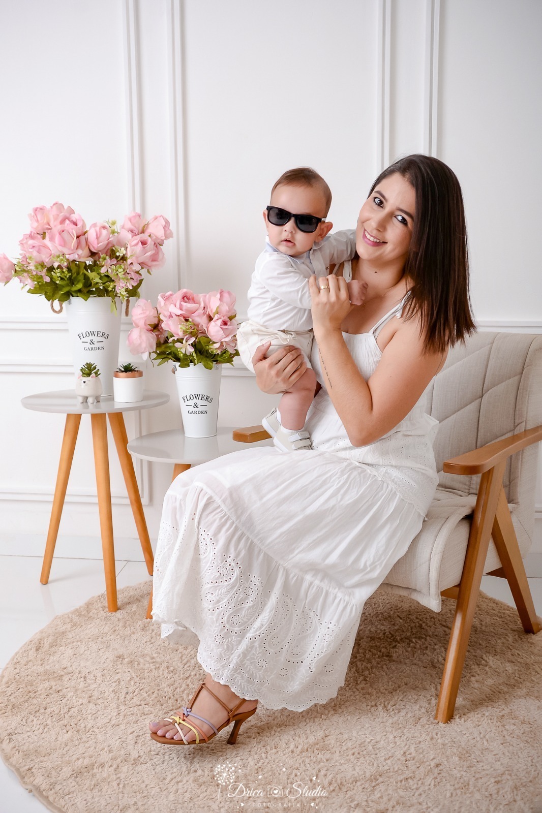 drica studio, ensaio fotografico, xinguara, dia das mães, mães segurando bebê nos braços, bebê usando óculos escuro, mesinha de canto, vaso branco com flores rosa, fundo branco com boiserie, poltrona de madeira e estofado cinza claro.
