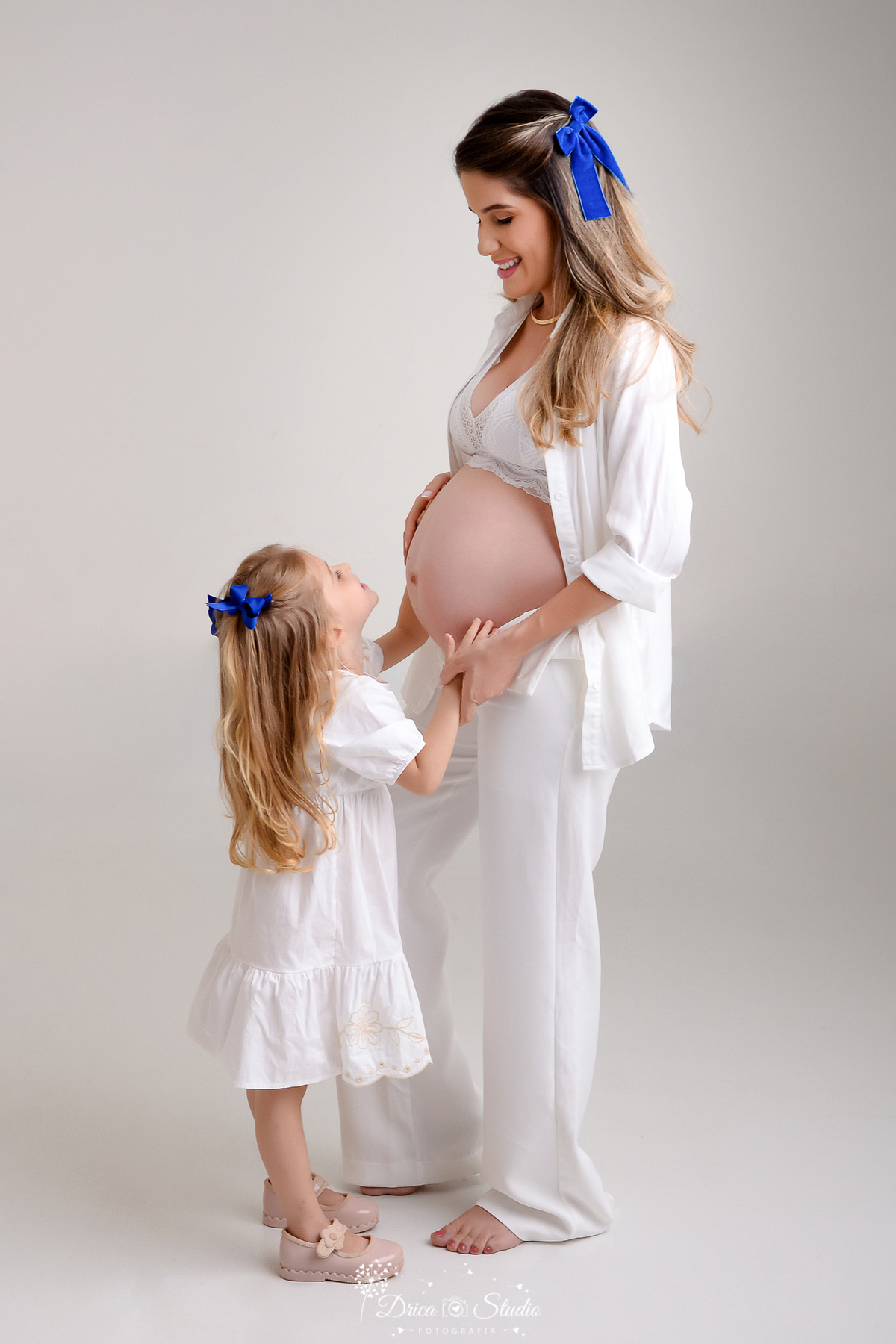 Drica Studio - ensaio fotográfico - gestante - xinguara - filha pequena acareciando barriga da mãe que está gestante - vestido branco - calça de alfaiataria branca - camisa e sutien branco - funco acizentado  - laço de cabelo azul