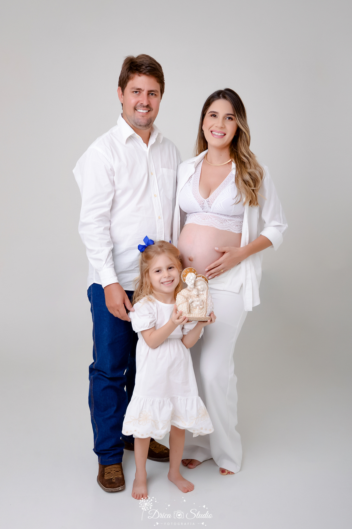 Drica Studio - ensaio fotográfico - gestante - xinguara - homem em pé ao lada da espora gestante e filha a frente segurando imagem de nossa senhora e o menino Jesus - camisa branca, - vestido branco - calça jeans - fundo cinza