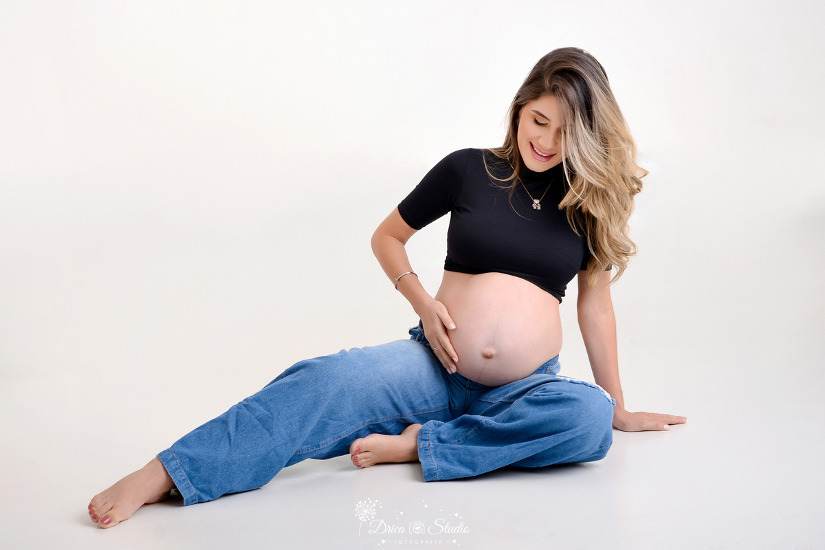 Drica Studio - ensaio fotográfico - gestante - xinguara - gestante sentada no chão com perna esticada para o lado, acareciando barriga - blusa preta - calça jeans - fundo branco - pingente menino e menina