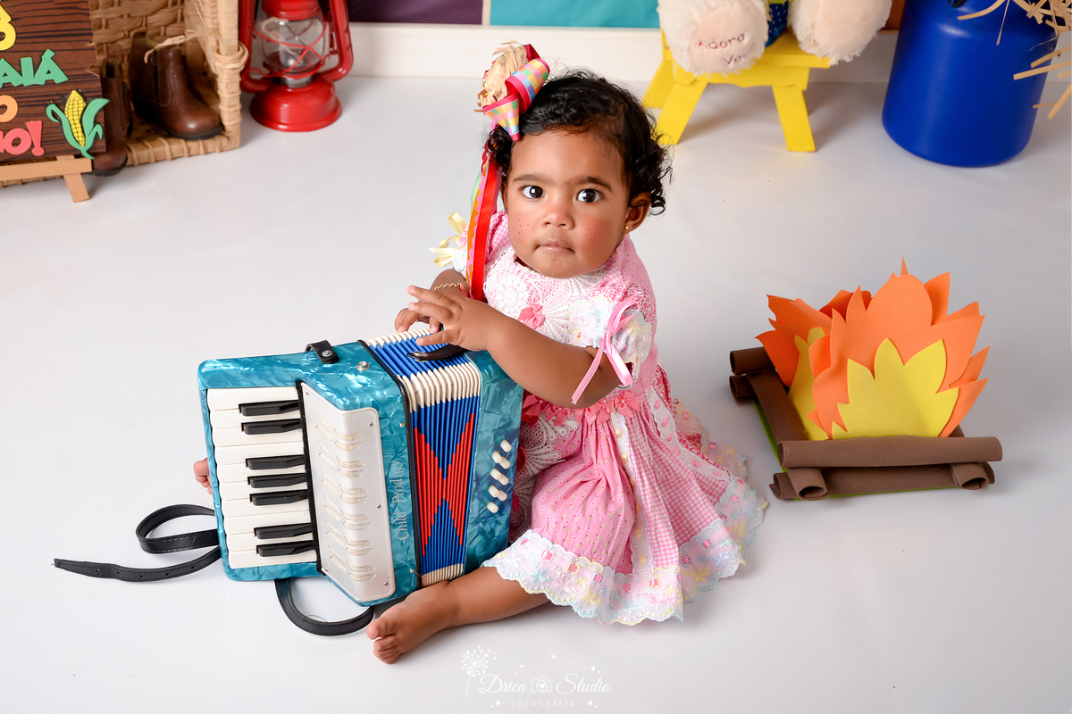 Drica studio - ensaio fotográfico infantil- xinguara - festa junina - menina sentada no chão segurando sanfona - fogueira - leiteira - lampião vermelho - vestido rosa - cadeira amarela