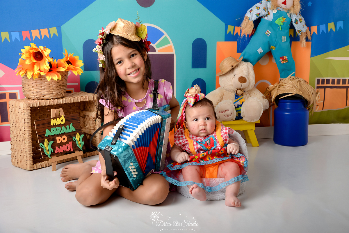 Drica studio - ensaio fotográfico infantil- xinguara - festa junina - menina segurando sanfona sentada ao lado da bebê sentada em poltrona - chapéu de palha decorado - girassol - latão de leite azul - espantalho - ursinho branco com chapéu de palha