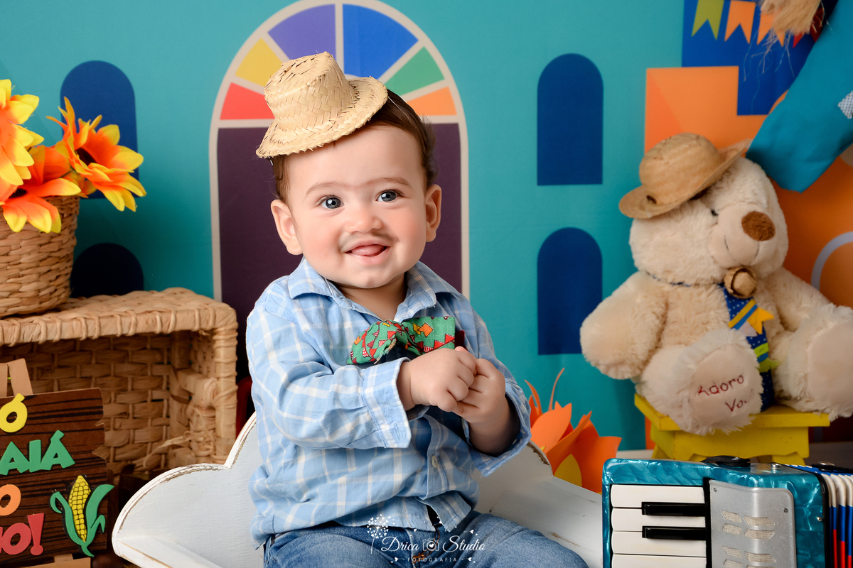 Drica studio - ensaio fotográfico infantil- xinguara - festa junina - bebê sentado em banquinho branco - camisa azul clara xadrez - calça jeans infantil - chapéu de palha - ursinho com chapéu de palha - sanfona