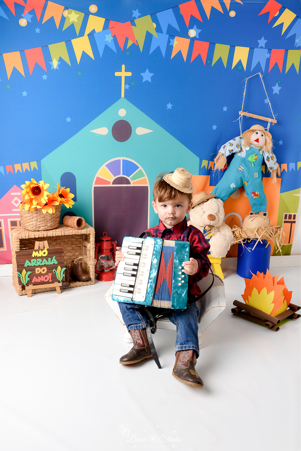 Drica studio - ensaio fotográfico infantil- xinguara - festa junina - menino sentado em banquinho branco com sanfona no colo - camisa infaltil xadrez vermelha com azul - espantalho - fogueiro - latão de leite azul - cesta de palha com girassol
