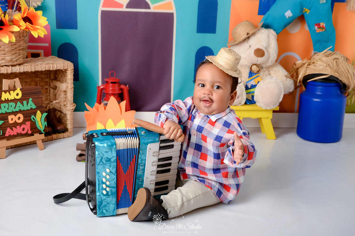 Drica studio - ensaio fotográfico infantil- xinguara - festa junina - bebê sentado no chão segurando sanfona azul - ursinho brannco com chapéu de palha - latão de leite azul - banquinho amarelo - cesto de palha - fundo tema junino