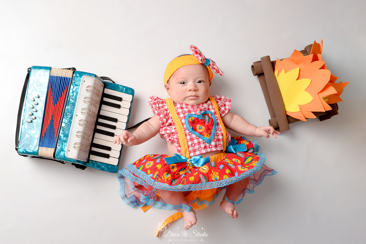 Drica studio - ensaio fotográfico infantil- xinguara - festa junina - bebê deitada no chão vestinda com vestindo junino - fogueira - sanfona azul e fundo branco