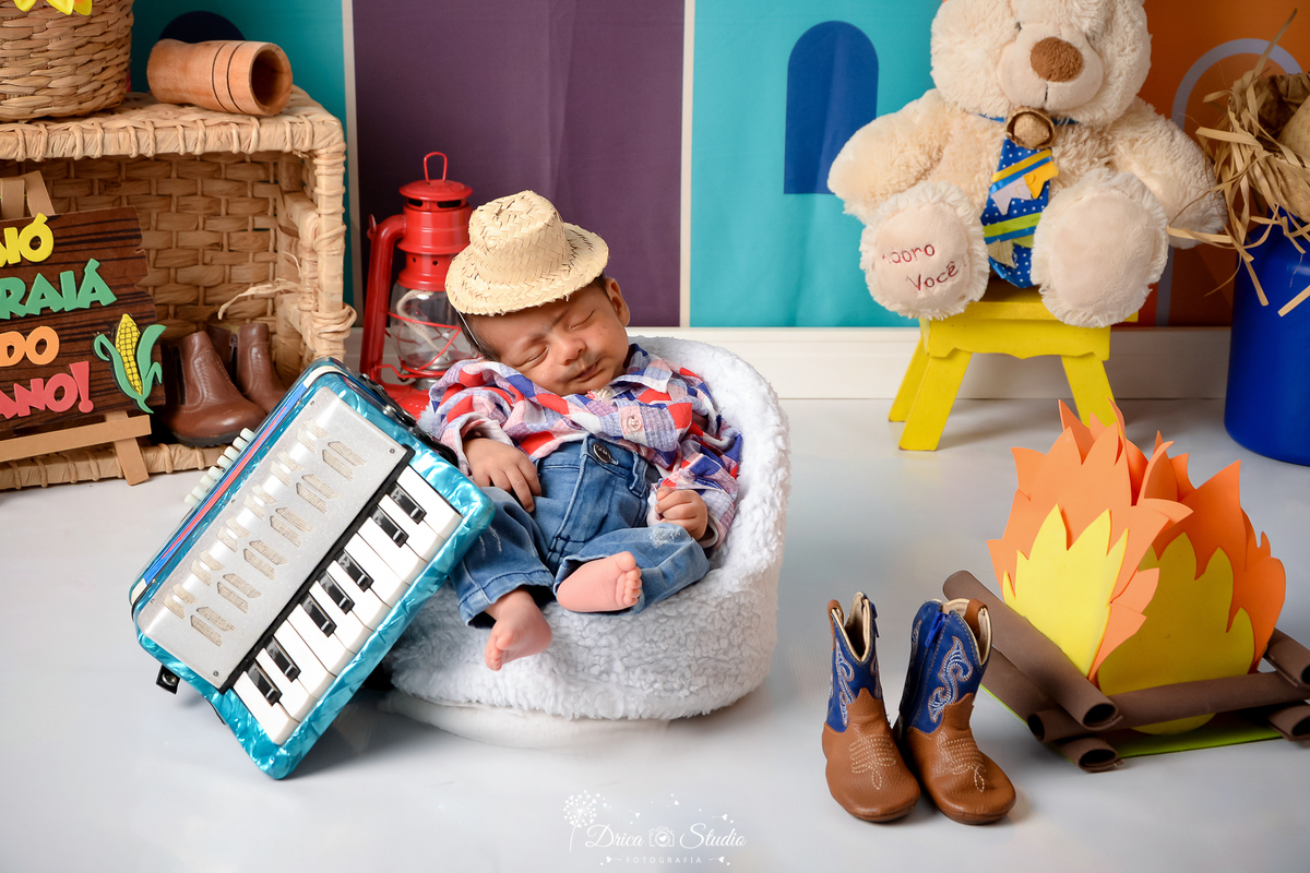 Drica studio - ensaio fotográfico infantil- xinguara - festa junina - bebê vestido de caipira dormindo sentado em poltrona branca - fogueira - sanfona azul - botas com detralhes azul - chapéu de palha - cesto de palha 