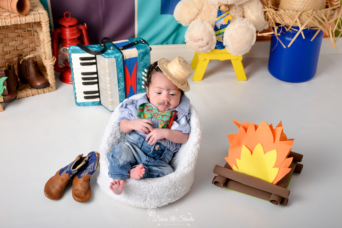 Drica studio - ensaio fotográfico infantil- xinguara - festa junina - bebê sentado em poltrona branca - fogueira - latão de leite azul - sanfona azul - cesta de palha - lampeão vermelho - urso branco com chapéu de palha