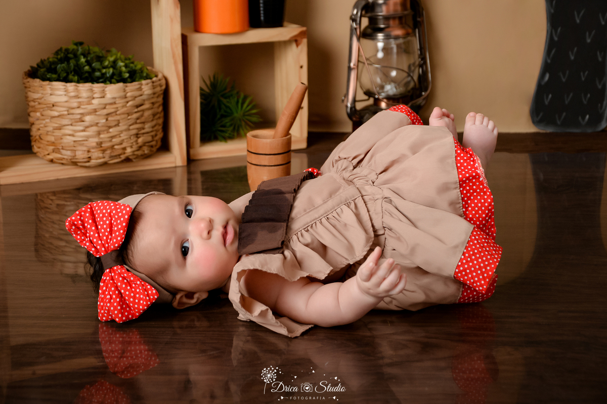 drica studio - ensaio fotografico infantil - tema sertão nordestino - bebê deitada no chão vetida de maria bonita - laço vermelho - cesto de palha com arbusto dentro - nicho de madeira - pilão de madeira - lampião antigo - fundo de cordel