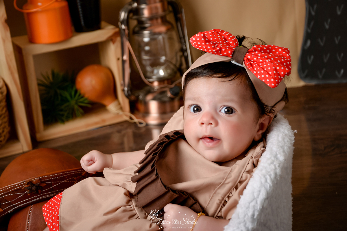 drica studio - ensaio fotografico infantil - tema sertão nordestino -  bebê sentado em poltrona branca - cabaça - lampião antigo - nicho de madeira - balde de leite laranja - laço vermelho - vestido bege - piso amadeirado