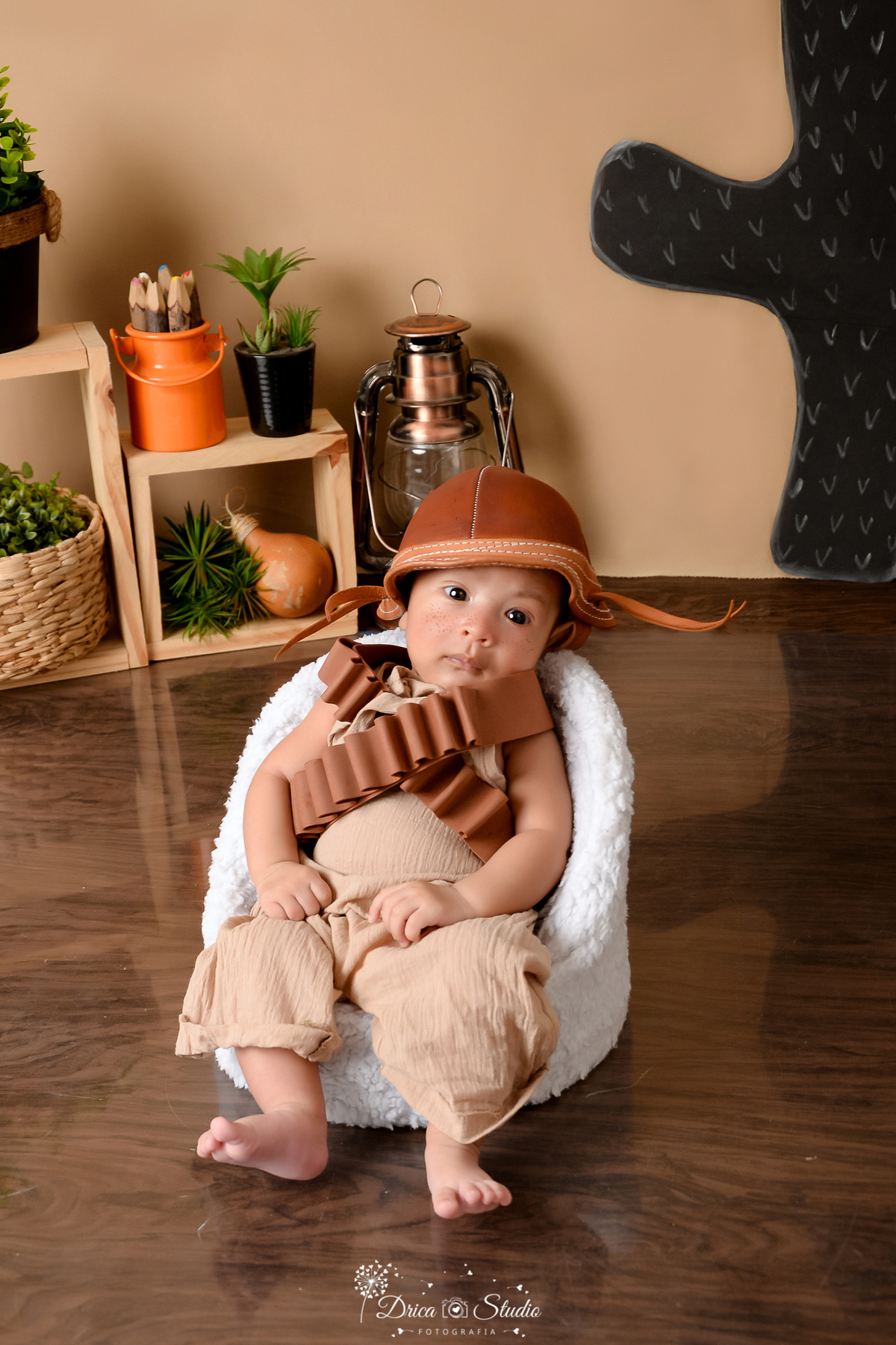 drica studio - ensaio fotografico infantil - tema sertão nordestino - bebê sentado em poltrona branca vestindo macaquinho bege - chapéu de cangaceiro - fundo de cordel - piso amadeirado - cesto de palha - cabaça