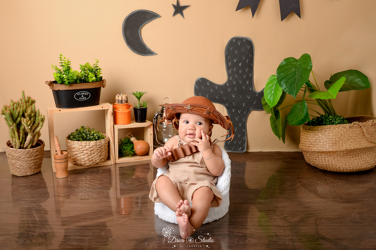 drica studio - ensaio fotografico infantil - tema sertão nordestino - bebê sentando em poltrona branca - chapéu de cordel - fundo de cordel - cestos de palha com arbustos dentro - latão de leite laranjado - nicho de madeira - cesto preto com arbustos 