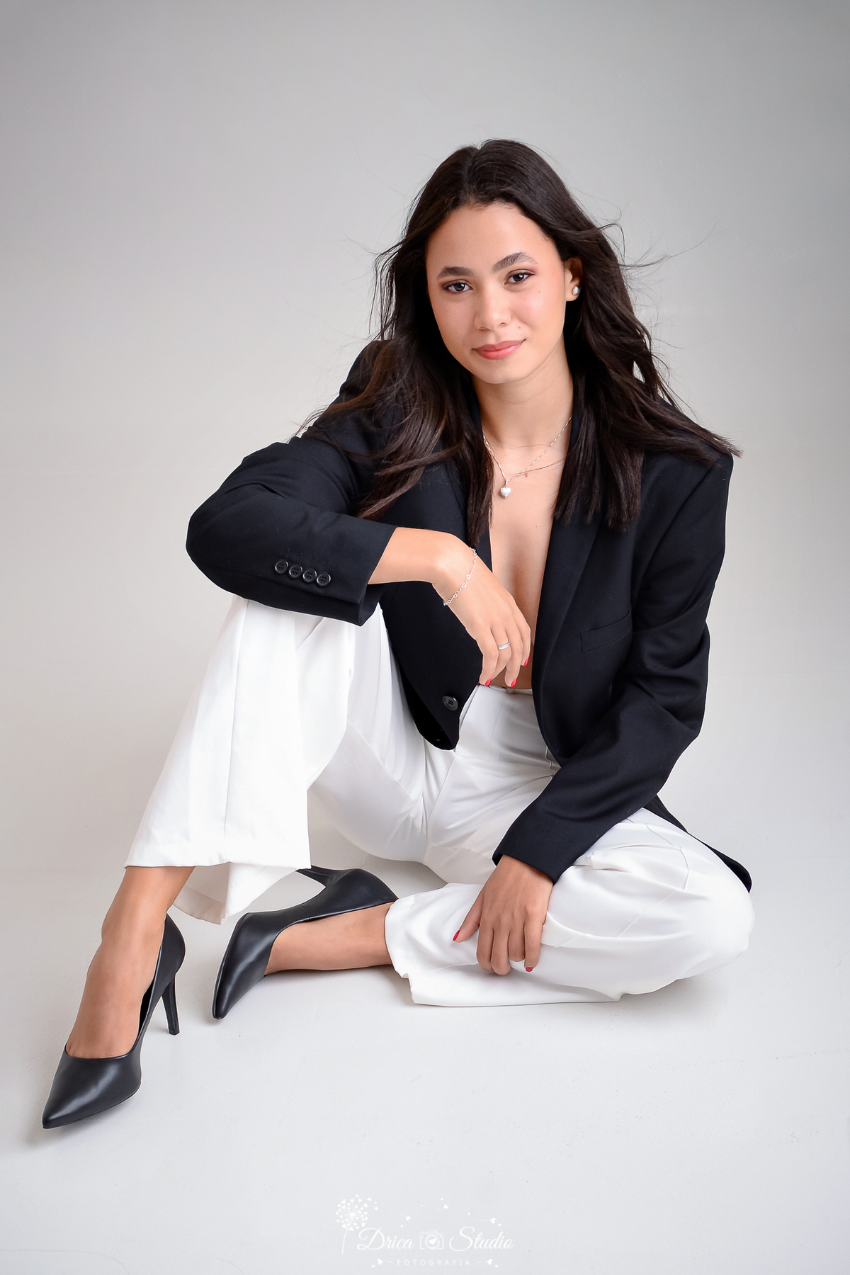 drica studio -ensaio fotográfico - aniversário 19 anos- mulher sentada no chão contra um fundo claro e neutro. Ela veste um blazer preto levemente aberto, sem blusa por baixo, combinando com uma calça branca de alfaiataria e sapatos de salto alto pretos. 