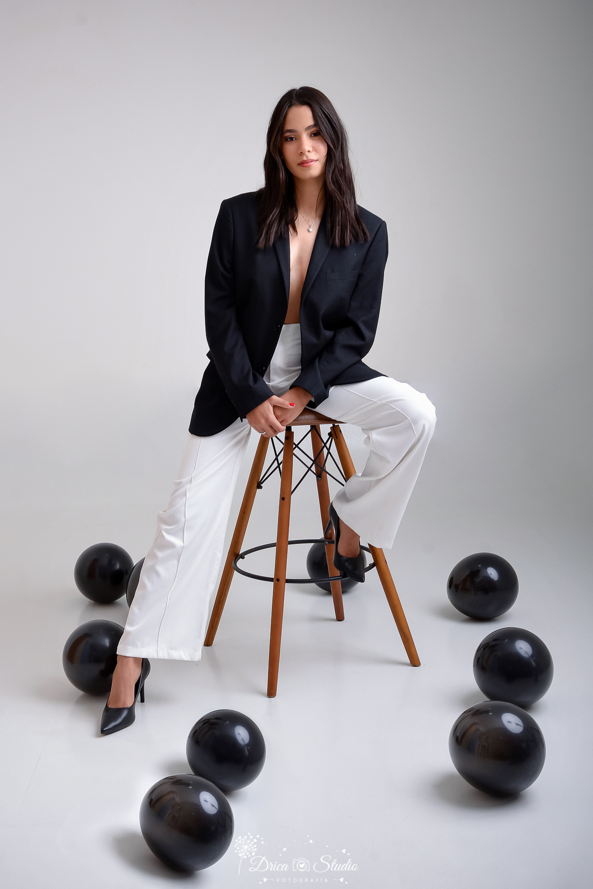drica studio - ensaio fotográfico - aniversário 19 anos - xinguara - mulher sentada em banco de madeira, vestindo blazer preto levemente aberto e calça branca, cabelos solto natural - balões preto   - fundo claro