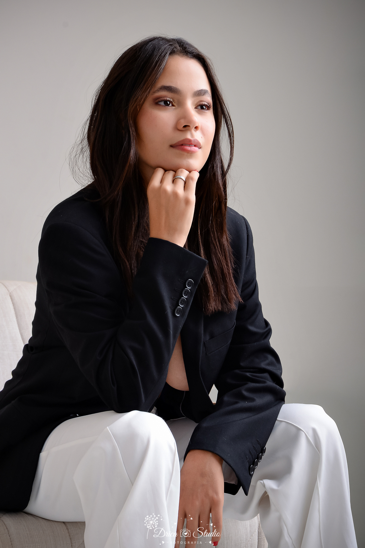 drica studio - ensaio fotográfico - aniversário 19 anos - xinguara - mulher sentada em uma poltrona clara, em um ambiente com fundo neutro, vestindo um blazer preto e uma calça branca, cabelos longo castanho escuro, levente ondulado e solto