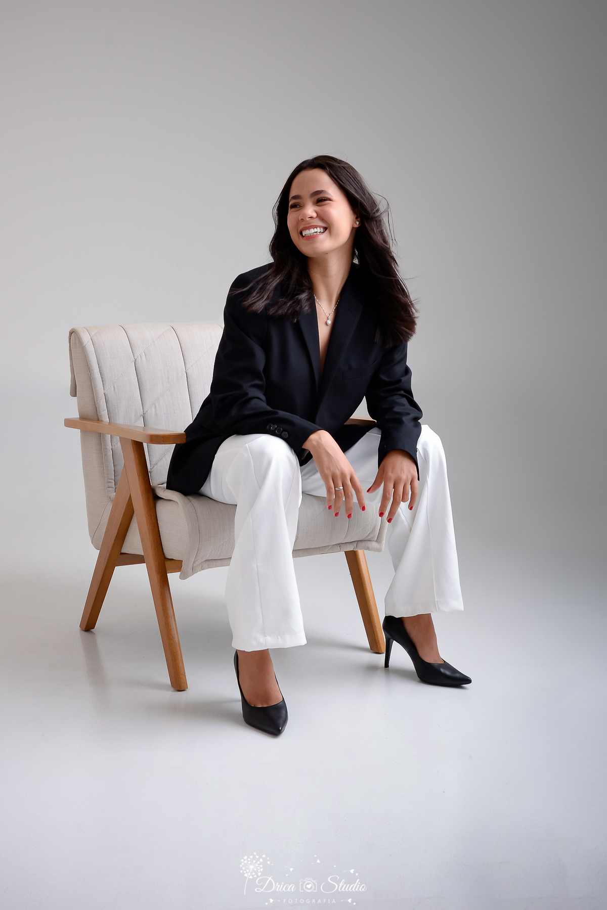 drica studio - ensaio fotográfico - aniversário 19 anos - xinguara -mulher sentada em uma poltrona clara, sorrindo de forma espontânea e alegre, com o cabelo solto e esvoaçante - usando blazer preto e calça branca - fundo claro