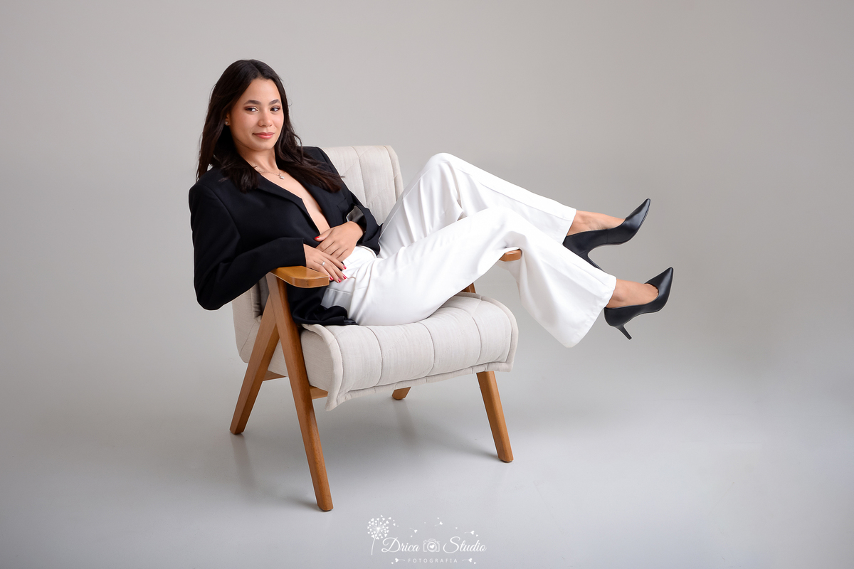 drica studio - ensaio fotográfico - aniversárlo 19 anos - xinguara -mulher reclinada em poltrona - usando blazer preto, calça branca e sapatos de salto alto pretos. Suas pernas estão esticadas, apoiadas sobre um dos braço da poltrona