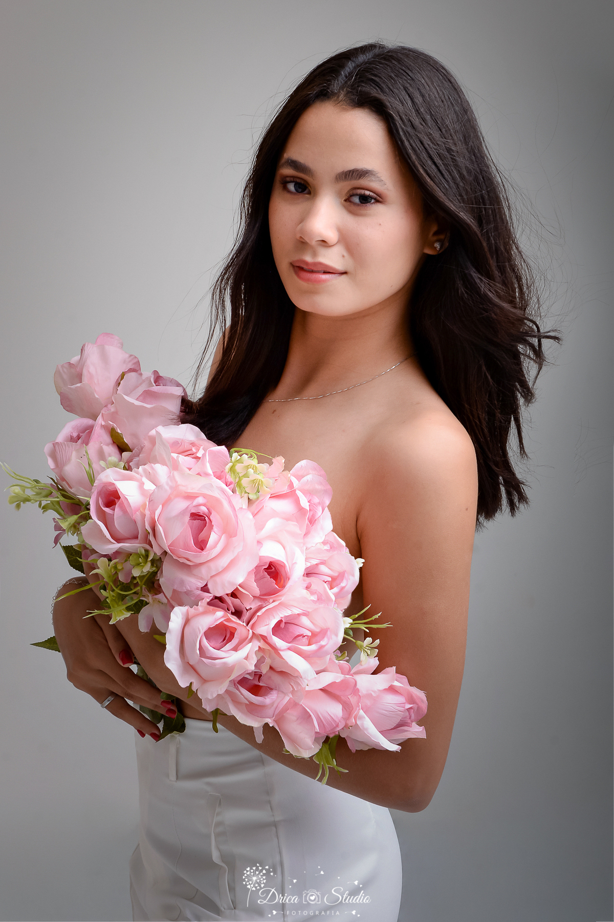 drica studio - ensaio fotográfico - aniversário 19 anos - xinguara -mulher segura junto ao corpo um volumoso buquê de rosas cor-de-rosa, Veste uma calça branca de cintura alta, e seu cabelo escuro está solto, com ondas suaves