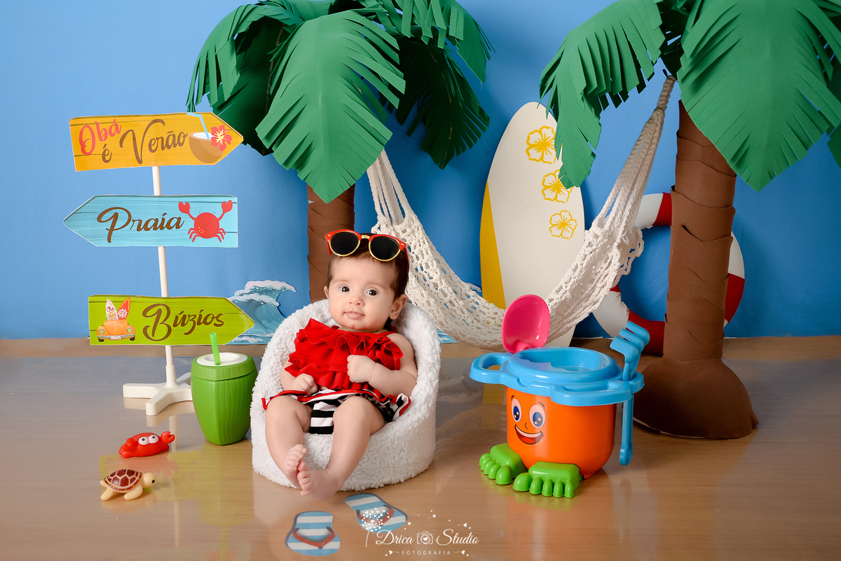 drica studio - ensaio fotográfico infantil - verão e praia - coqueiros - rede branca - prancha de sulf - bebê vestindo top vermelho e short listrado - baldinho de brinquedo laranja - bebê sentado em poltrona - fundo azul emitando o céu.