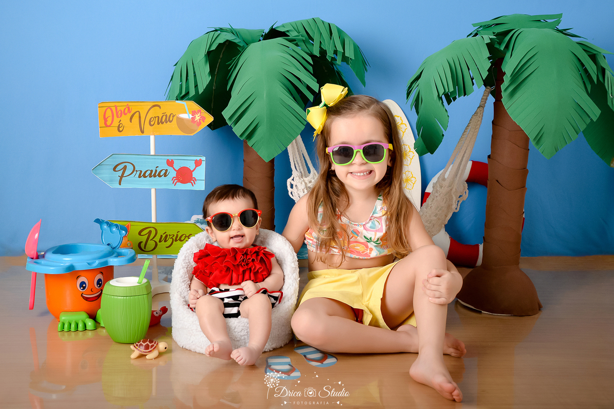 drica studio - ensaio fotográfico infantil - verão e praia - coqueiros - bebê vestindo top vermelho e short listrado e irmã mais velha vestindo top floral com short amarelo - fundo azul - coco verde com canudo - óculos de sol