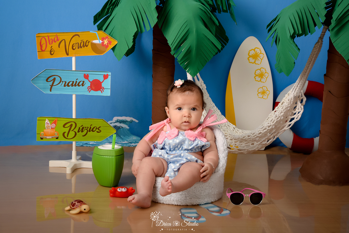 drica studio - ensaio fotográfico infantil - verão e praia - coqueiros - prancha branca com flores amarela - bebê sentada em poltrona branca vestindo biquine lilás com rosa - fundo azul - tartaruga e carangueijo de brinquedo - óculo de sol rosa