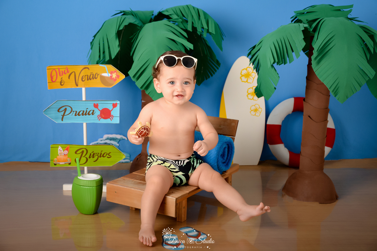 drica studio - ensaio fotográfico infantil - verão e praia - coqueiros - bebê vestindo sunga de praia sentado em espreguiçadeira usando óculos de sol sobre a cabeça - prancha branca com flores amarela - fundo azul 
