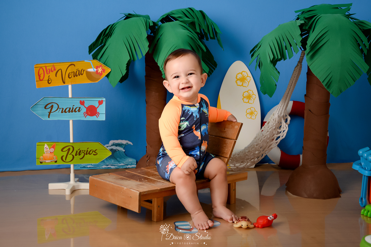 drica studio - ensaio fotográfico infantil - verão e praia - coqueiros - bebê sorridente, vestindo roupas de sulfista, sentado em espreguiçadeira de madeira - carangueijo vermelho - prancha de sulf branca - rede branca - 