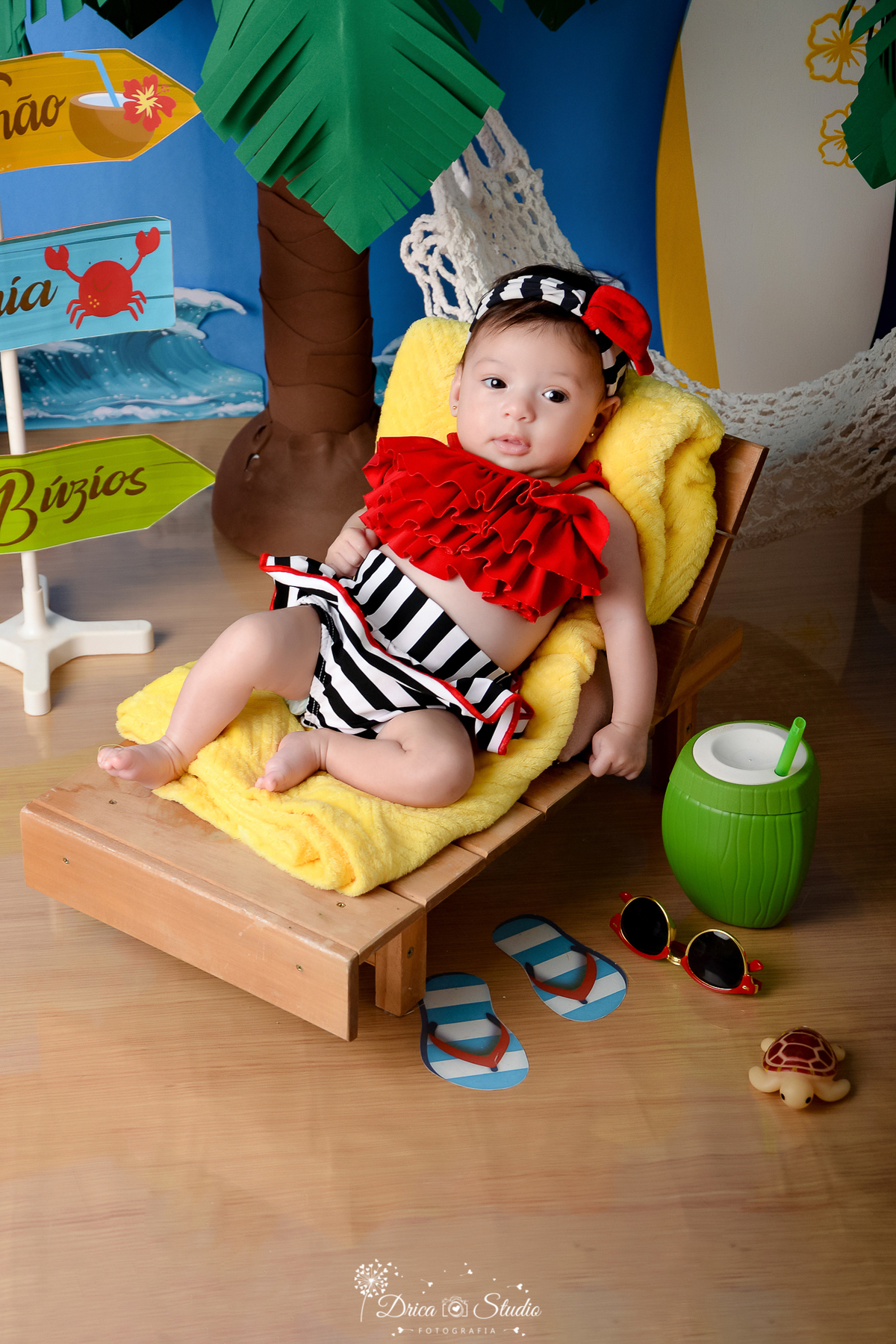 drica studio - ensaio fotográfico infantil - verão e praia - bebê vistindo top vermelho e short com listras branca e preta deitada em espreguiçadeira de praia - coco verde com canudo -  óculos de sol - toalha amarela