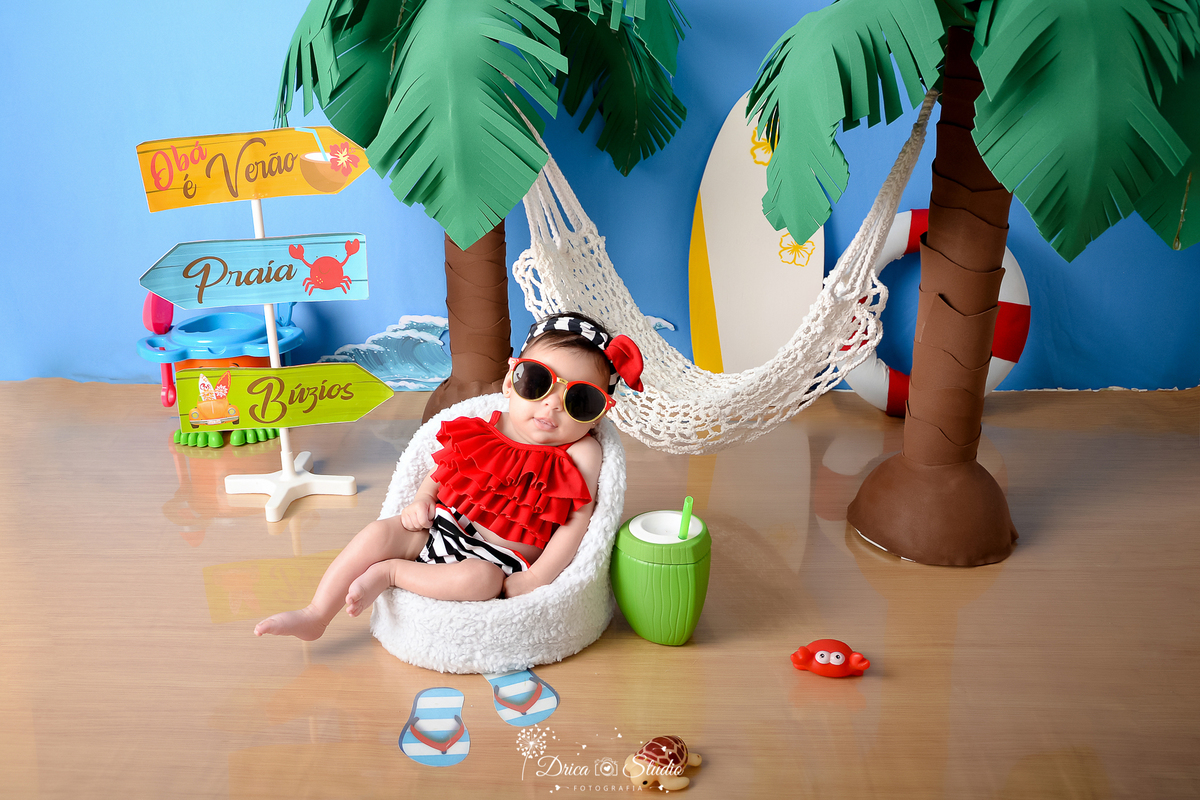drica studio - ensaio fotográfico infantil - verão e praia - bebê vestindo top vermelho e short com listras preta e branca sentada em poltrona branca - rede branca - coqueiros - prancha de sulf - placas de orientação - coco verde com canudo 