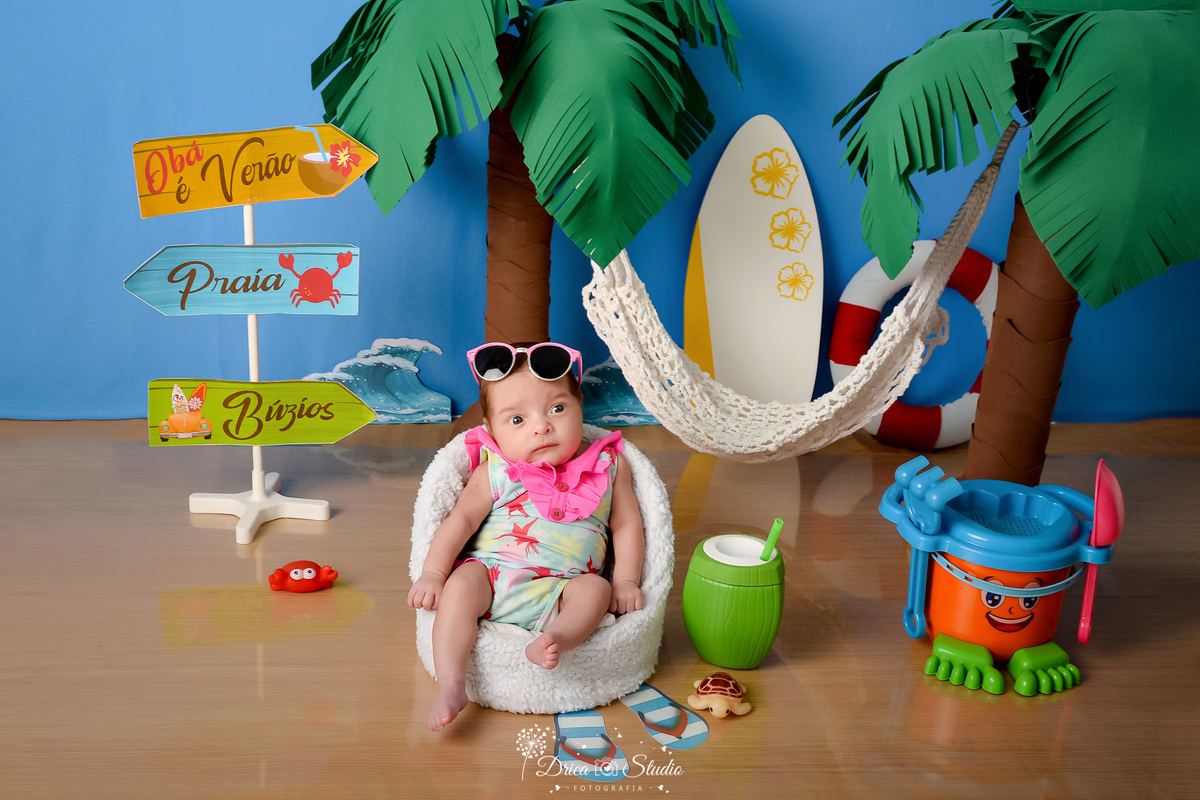 drica studio - ensaio fotográfico infantil - verão e praia - bebê vestindo maiô florado com babado rosa sentada em poltrona branca - baldinho de brinquedos laranja com verde e azul - conqueiros - prancha de sulf - coco verde com canudo