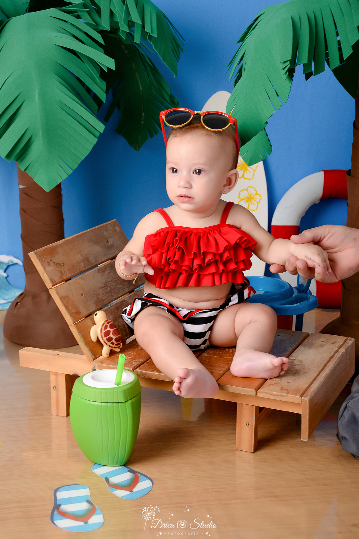 drica studio - ensaio fotográfico infantil - verão e praia - bebê sentada em espreguiçadeira vestindo top com babados vermelho e short com listras branca com preto - boia salva vida - coqueiro - fundo azul