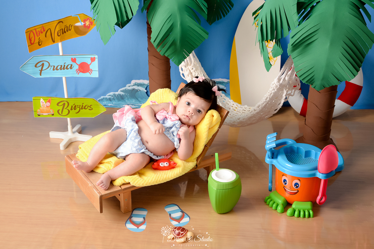 drica studio - ensaio fotográfico infantil - verão e praia - bebê vestindo biquini azul com babado rosa - baldinho de brinquedo laranja com azul e verde - coqueiros - pranca branca com flores amarela - rede branca - fundo azul 