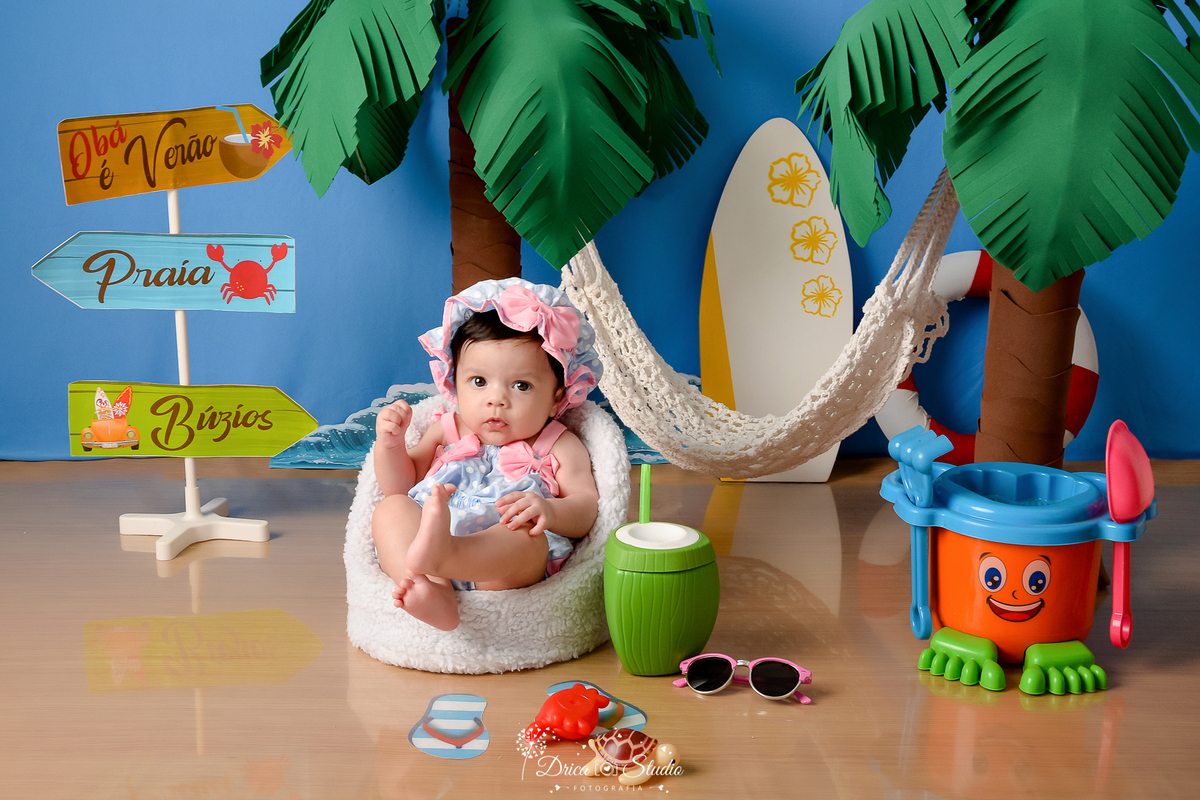drica studio - ensaio fotográfico infantil - verão e praia - bebê vestindo biquini azul com babado rosa sentada em poltrona branca - coco verde com canudo - coqueiros - rede branca - fundo azul - prancha branca com flores amarelas - óculos de sol 