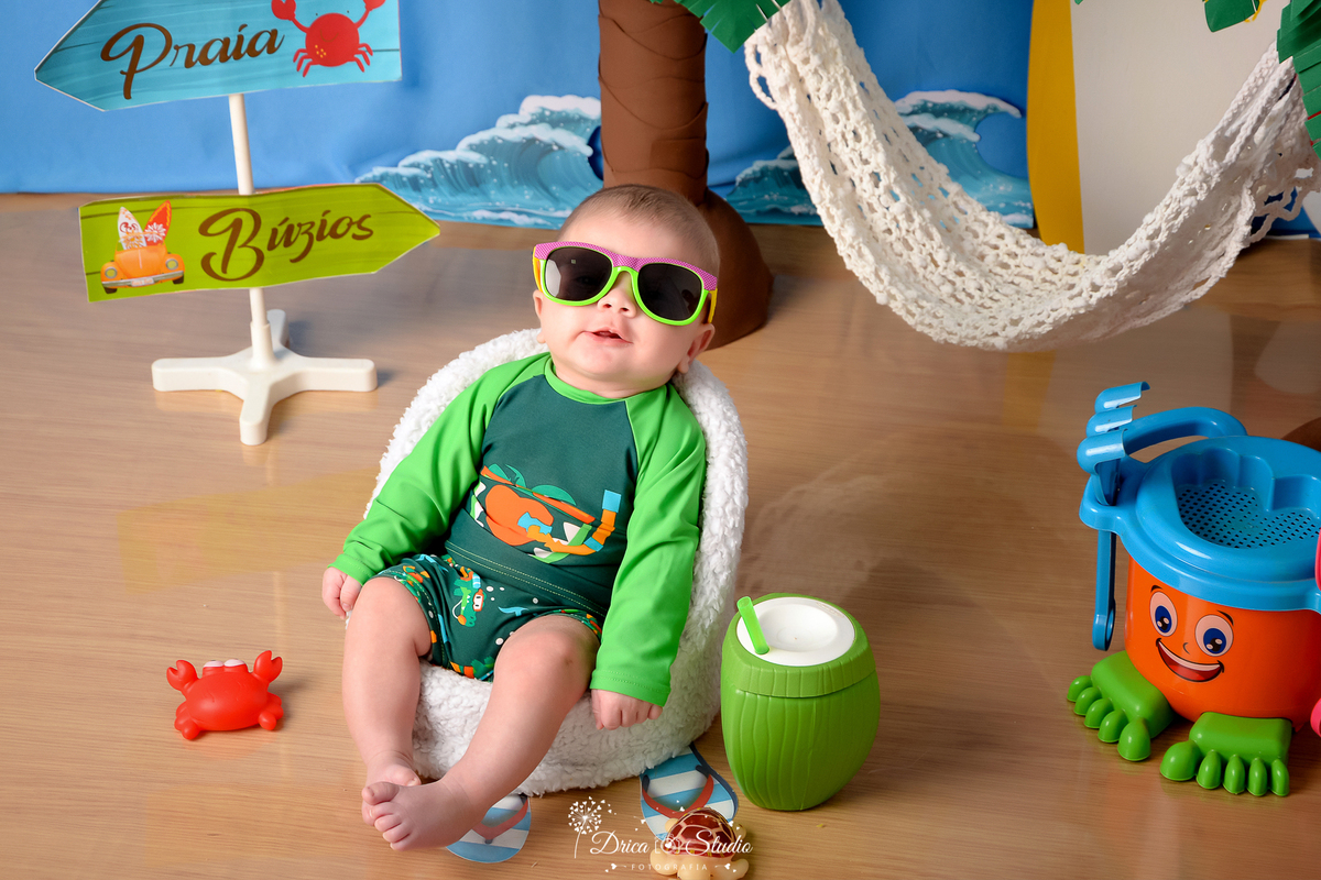 drica studio - ensaio fotográfico infantil - verão e praia - bebê vestindo roupa termica de banho sentado em poltrona branca - baldinha de brinquedo laranja com azul e verde - coco verde com canudo - rede branca - fundo azul - plaquinhas de indicação