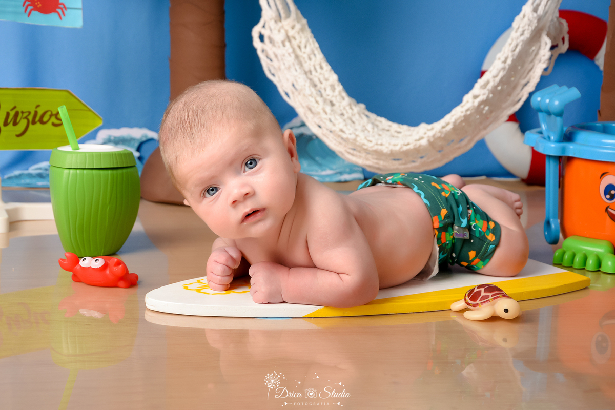 drica studio - ensaio fotográfico infantil - verão e praia - bebê vestindo sunga verde estampada deitado de brurços sobre prancha de sulf - coco verde com canudo - carangueijo e tartaruga de brinquedo - funco azul - rede branca - piso emitando areia