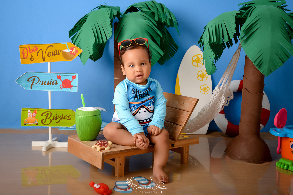 drica studio - ensaio fotográfico infantil - verão e praia - bebê vestindo camisa termica azul clara sentado em espreguiçadeira e com óculos de sol sobre a cabeça - coqueiros - coco verde com canudo - prancha de sulf branca cm flores amarelas - boia