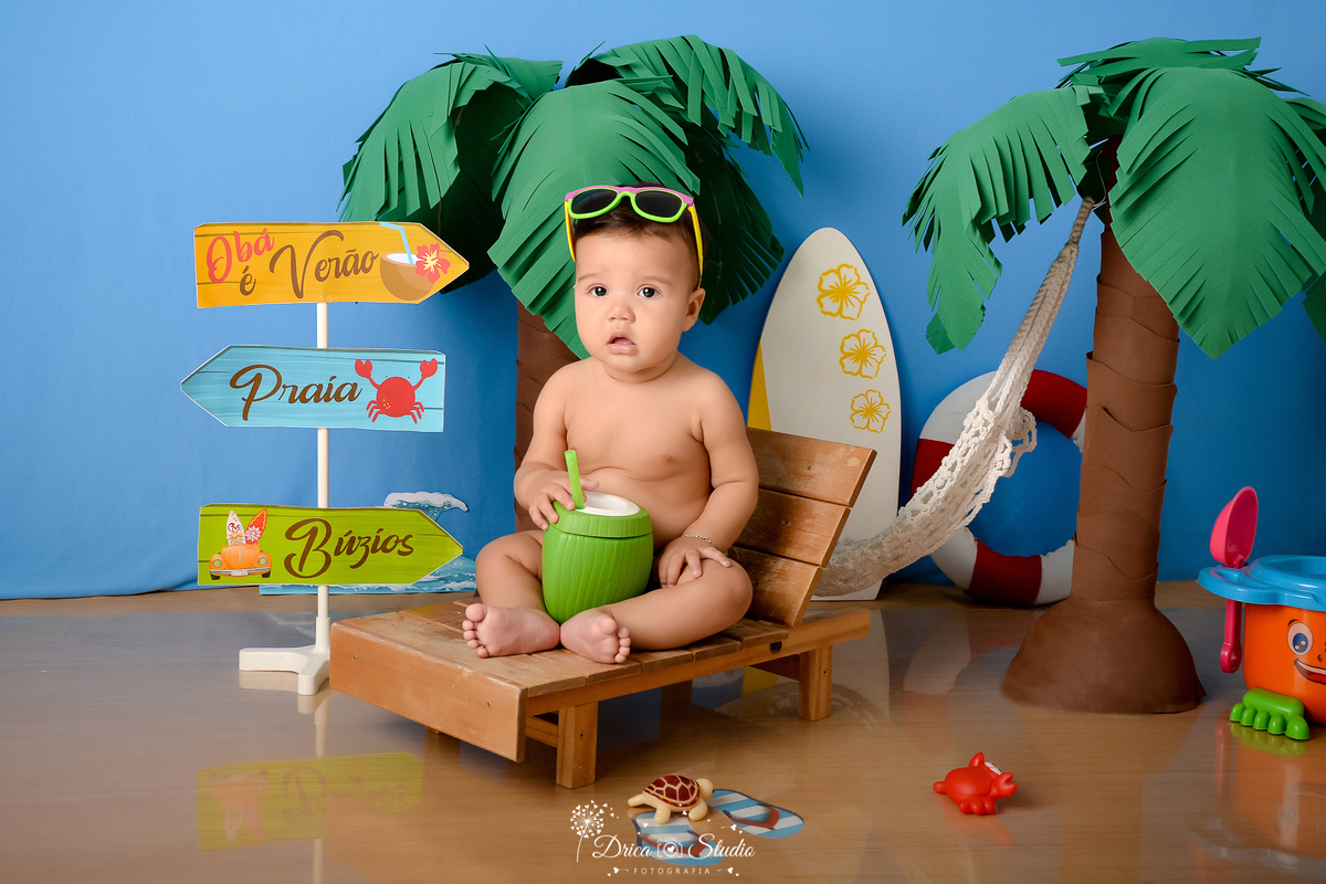 drica studio - ensaio fotográfico infantil - verão e praia - bebê sentado em espreguiçadeira segurando um coco verde com canudo e usando óculos de sol sobre a cabeça - coqueira - boia salva vida - fundo azul - prancha branca com flores amarela - plaquinha
