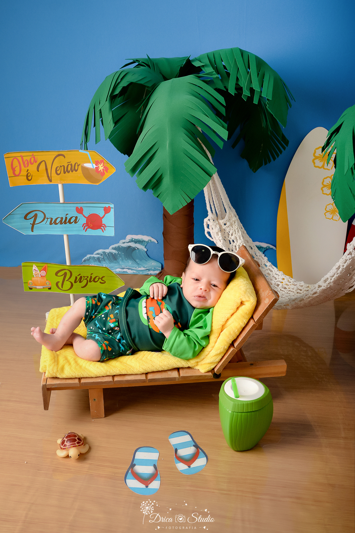 drica studio - ensaio fotográfico infantil - verão e praia - bebê vestindo roupa termica de praia deitado sobre toalha amarela em espreguiçadeira - rede branca - fundo azul - ondas do mar - prancha branca com detalher amarelo - coqueiros - óculos de sol