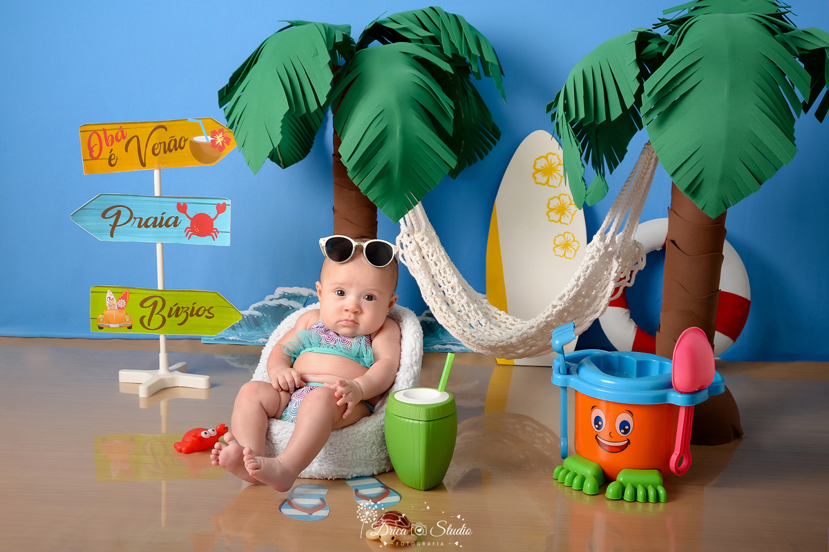 drica studio - ensaio fotográfico infantil - verão e praia - bebê sentada poltrona branca, vestindo biquini estampado com babado azul, com óculos de sol sobre a cabeça - coqueiros - prancha de sulf branca com flores amarelas - plaquinhas de indicação 