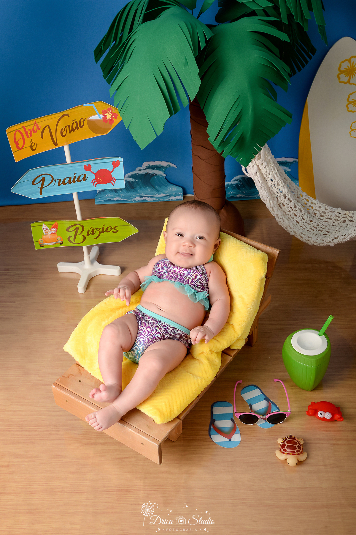 drica studio - ensaio fotográfico infantil - verão e praia - bebê deitada sobre manta amarela em espreguiçadeira, usando biquini estampado com babado azul - coco verde com canudo - plaquinhas de indicação - ondas do mar - rede branca - coqueiro 