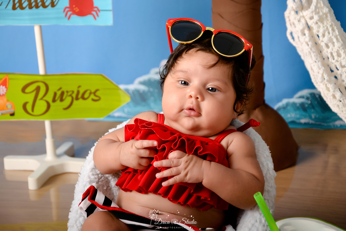 drica studio - ensaio fotográfico infantil - verão e praia - bebê vestindo top vermelho e short com listras branca e preta, usando óculos de sol sobre a cabeça - fundo azul - ondas do mar - rede branca - plaquinhas de indicação - piso emitando areia