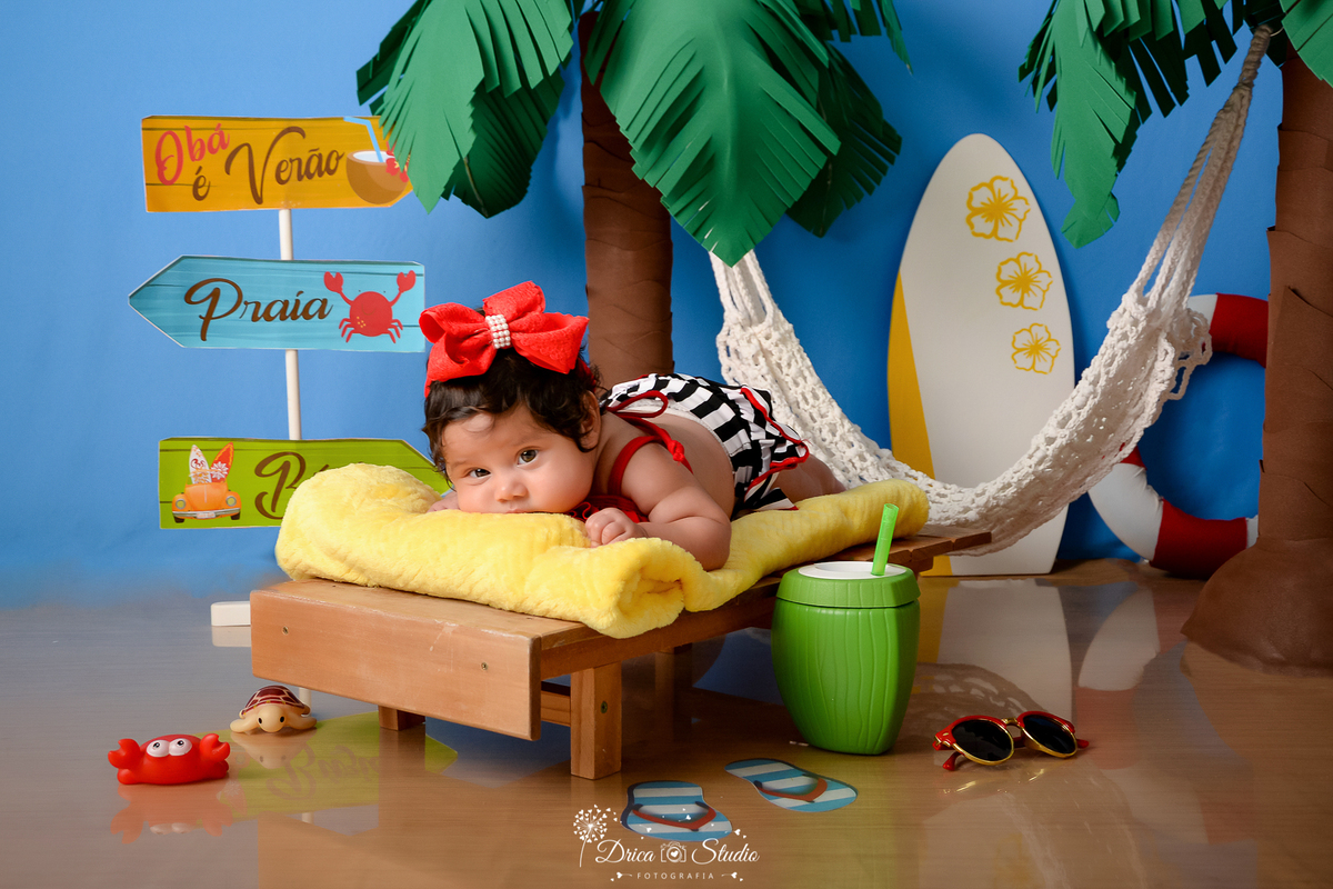 drica studio - ensaio fotográfico infantil - verão e praia - bebê vestindo short com listras branca com preto, usando laço vermelho no cabelo deitada de bruços em espreguiçadeira - prancha branca com flores amarela - coqueiros - fundo azul - rede branca